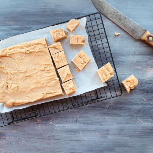 Block of delicious, homemade peanut butter fudge being cut into squares over a textured wood table background with old knife. Image shot from above.