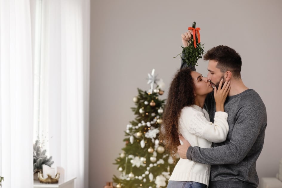 Happy couple kissing under mistletoe bunch in room decorated for Christmas.