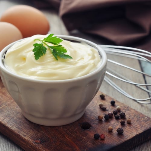 Homemade mayonnaise on wooden board with eggs in background.
