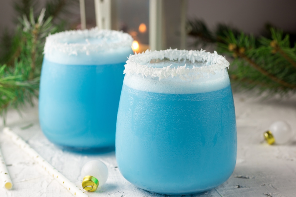 Blue curacao Christmas Cocktail, garnished with coconut on Christmas decorated holiday table with Christmas ornaments. Holiday cocktails with ice Blue Curacao, coconut cream, vodka and pineapple juice.