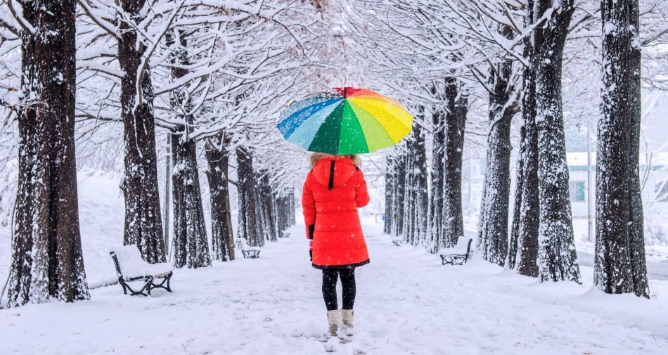 A woman in a red raincoat holding a rainbow colored umbrella while walking in a snowy forest.