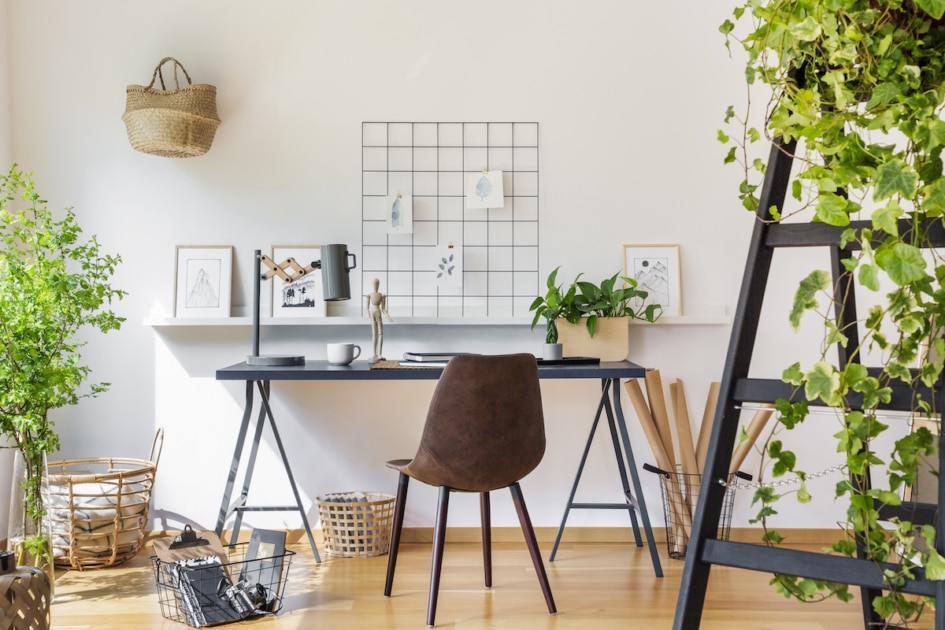 Brown chair at desk in white boho home office interior with plants and posters.