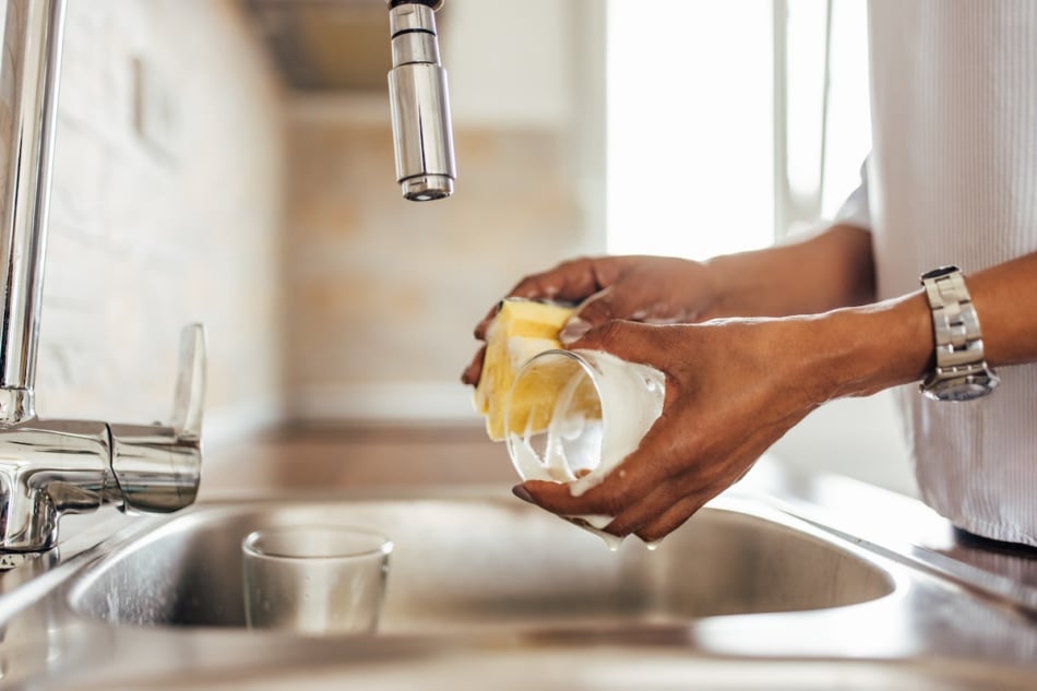 Person using sink and cleaning dishes, holding glass and sponge.