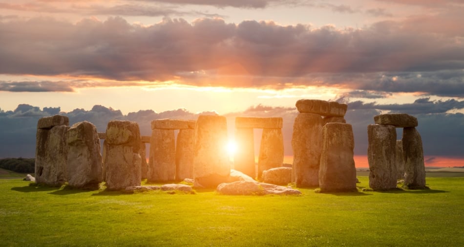 Stonehenge under a cloudy sky with sun shining behind it.