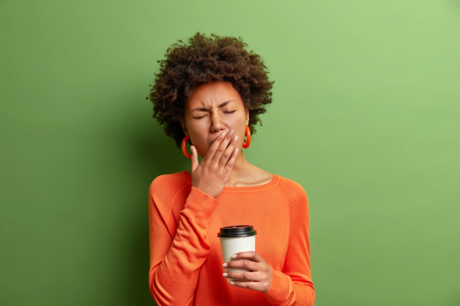 Photo of beautiful tired woman yawns and being sleepy after awakening early in morning holds disposable cup of coffee dressed in casual jumper isolated on vivid green background. Exhausted woman