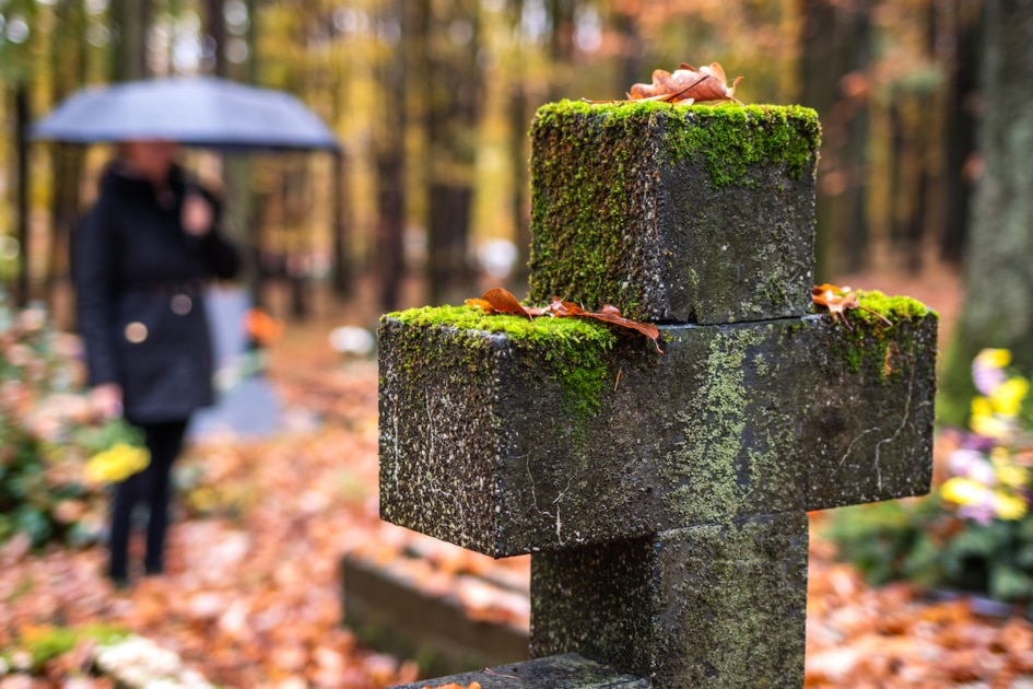 Religious cross in cemetery. Mourning woman in black standing next to tombstone in rain. Silent memory for dead relatives