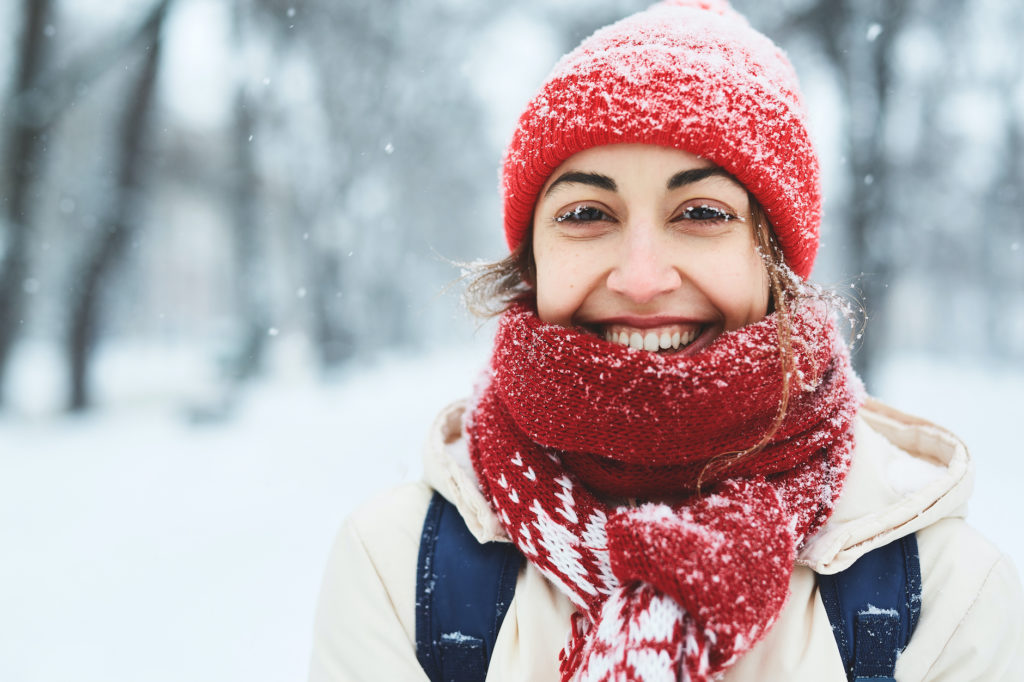 Happy cheerful smiling woman in warm clothes, red knitted cap, scarf and mittens walking on the snowy street after blizzard in city.
