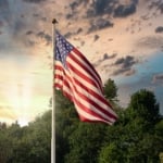 American flag waving on a pole with tress and sun shining in background.