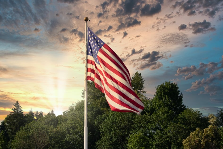 American flag waving on a pole with tress and sun shining in background.