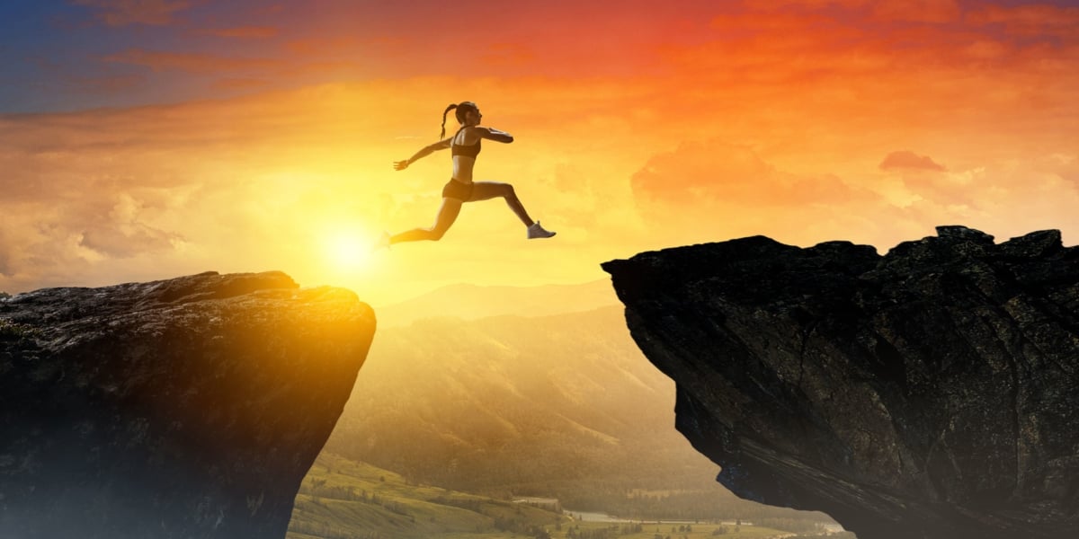 Female athlete leaping from one cliff to another with sun setting in background.