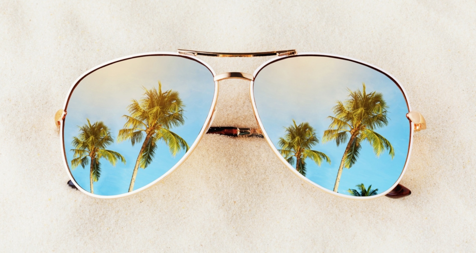 Sunglasses on sand with lenses showing images of palm trees and blue skies.