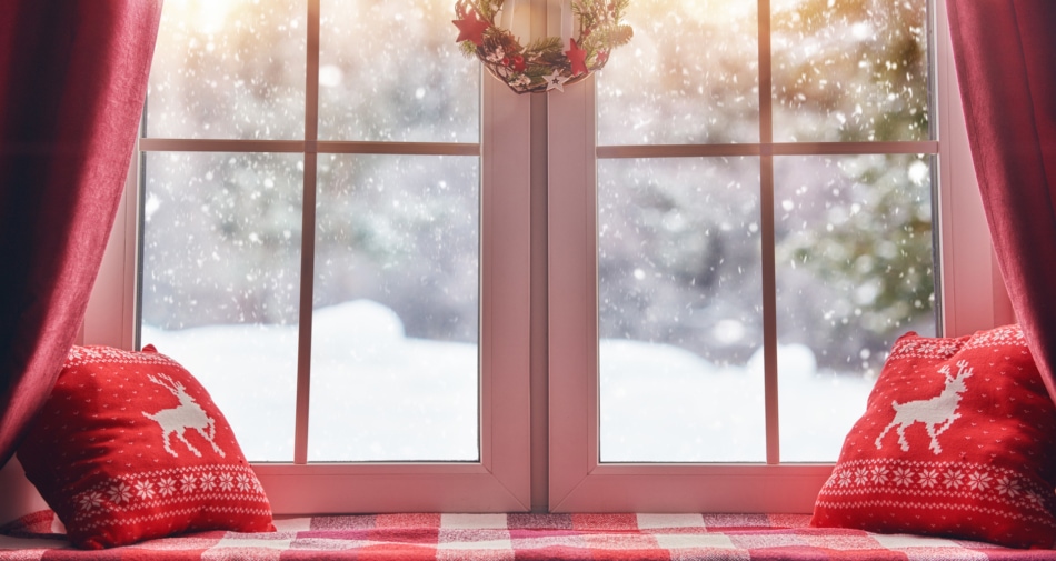 Two red Christmas themed pillows adorning a window seat, with snow falling in the background.