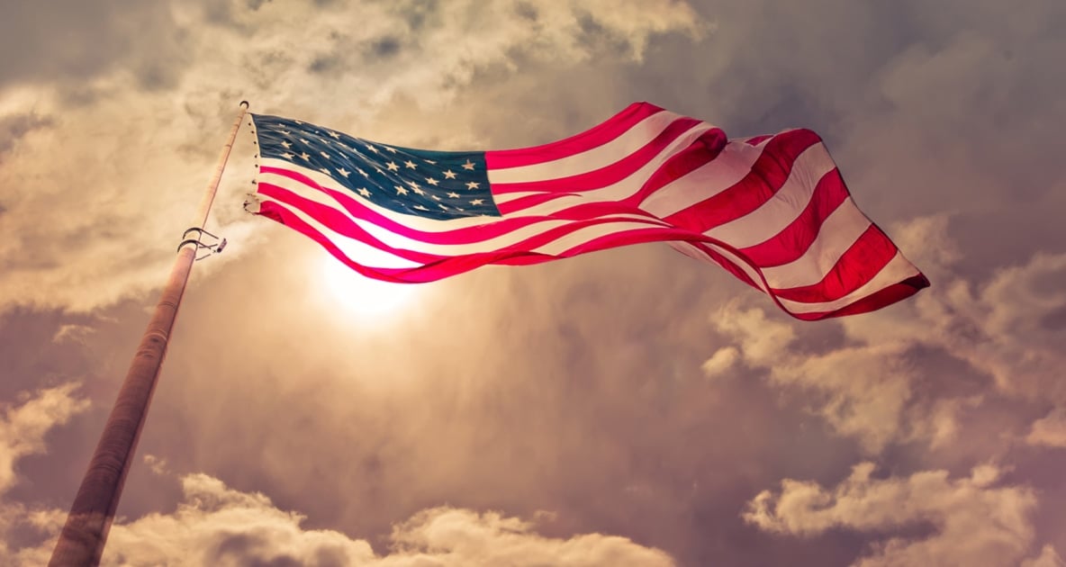 American Flag towering in sky with sun and clouds overhead.