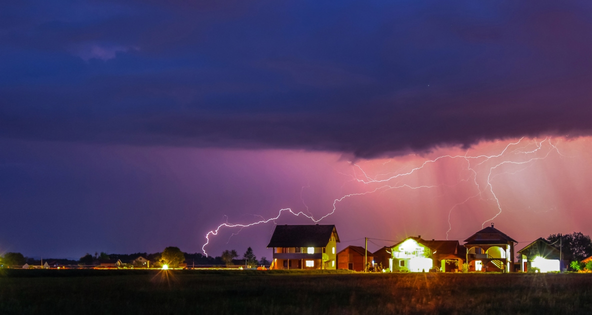 Thunderstorms with a purplish hue striking lightning over a well lit neighborhood.