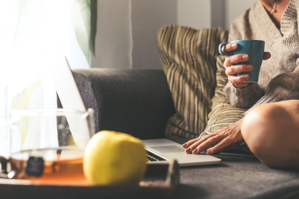 Woman sitting on a couch in the living room at home using laptop in a winter morning. Girl working on the sofa in cozy soft comfortable sweater, having natural breakfast with tea coffee and fruits