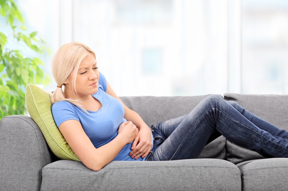 Blond woman with a painful expression sitting on a grey sofa at home with her hands placed on her stomach.