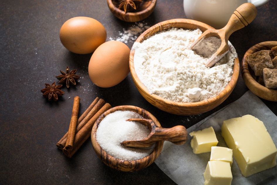 Arranged baking ingredients for cooking, including flour, eggs, butter, sugar, milk and spices.