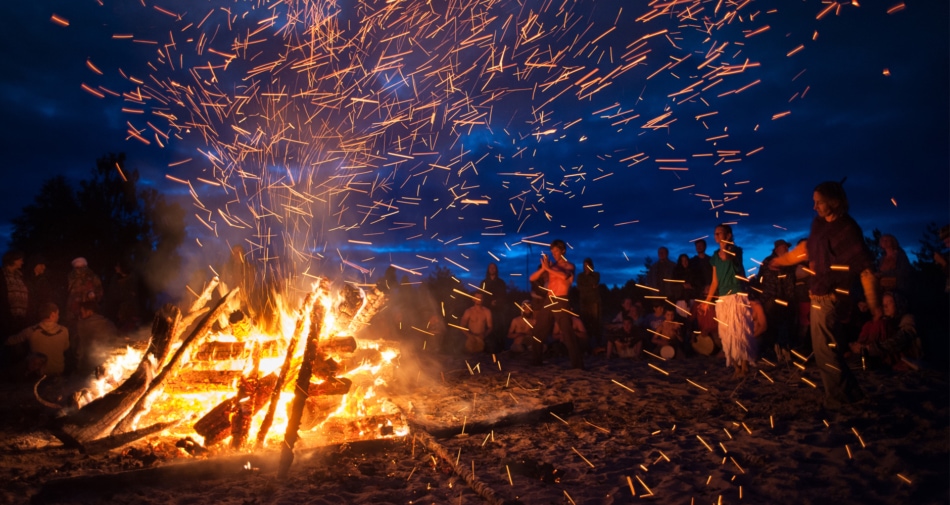 Group of people dancing in front of a bonfire on a beach at night.