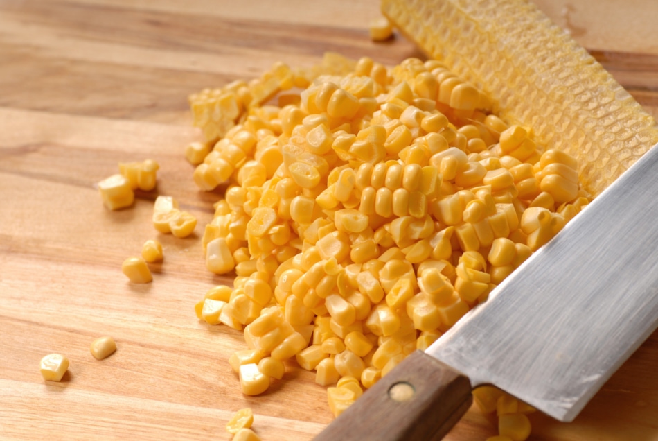 Corn kernels cut off the cob with a sharp knife on a cutting board.