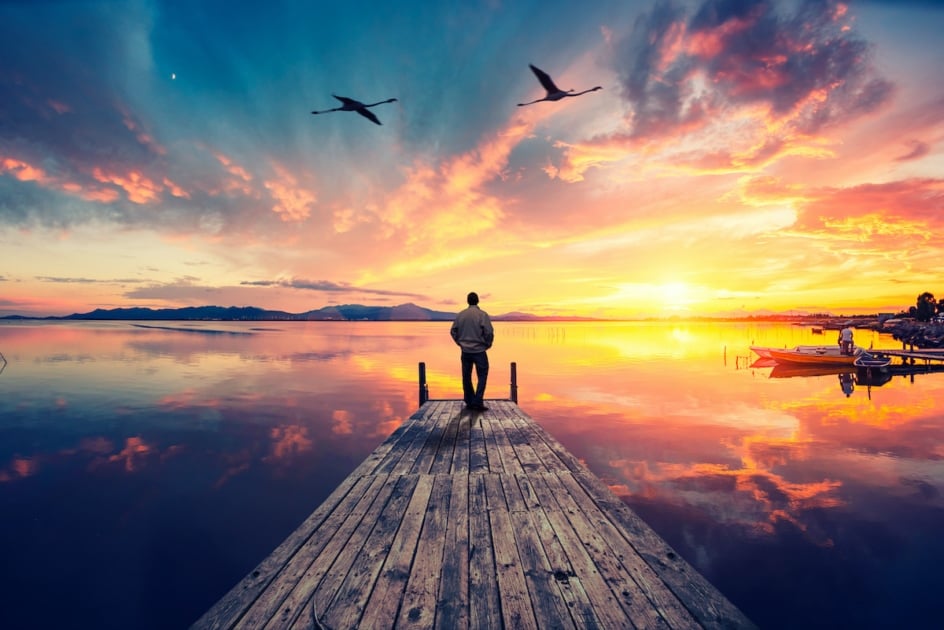 Lonely man on wooden pier contemplating a colorful sunset reflected on the sea with two flamingos flying high.