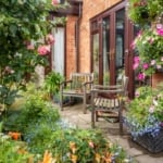 Two weathered wooden chairs sit on a stone patio surrounded by blooming petunias and lush garden greenery.