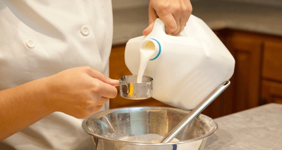 Baker pouring milk into a measuring cup over a bow of flour.