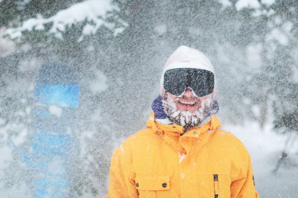 Skier's or snowboarder portrait. Bearded man in winter hat and black goggles at snow blizzard in the forest. Happy and smiling snowboarder with snow on his face.