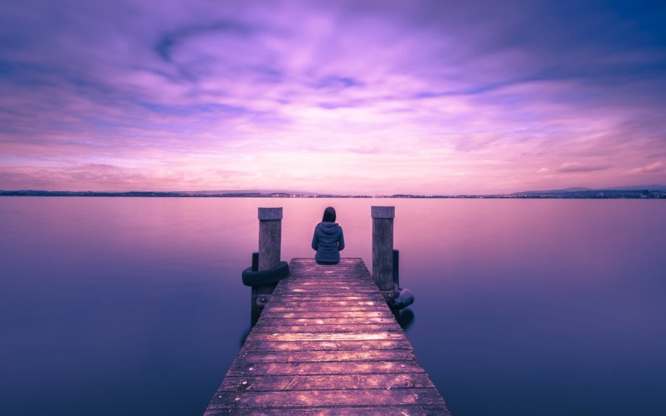 One woman sits on a wooden pier. Cloudy above the lake.