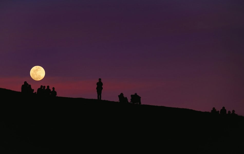 People watching the full moon from a hill at dusk.