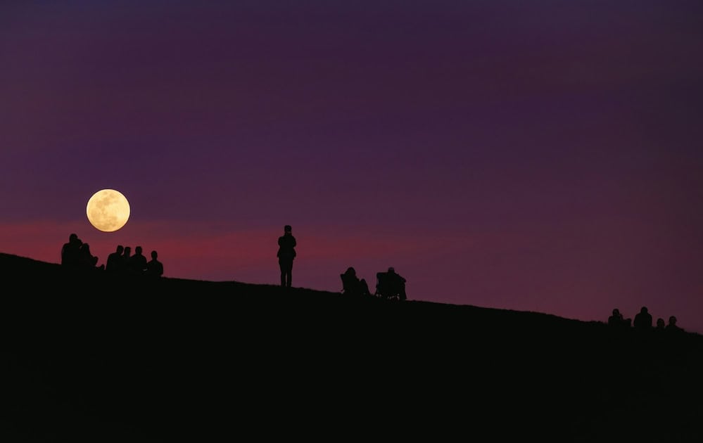 People watching the full moon from a hill at dusk.