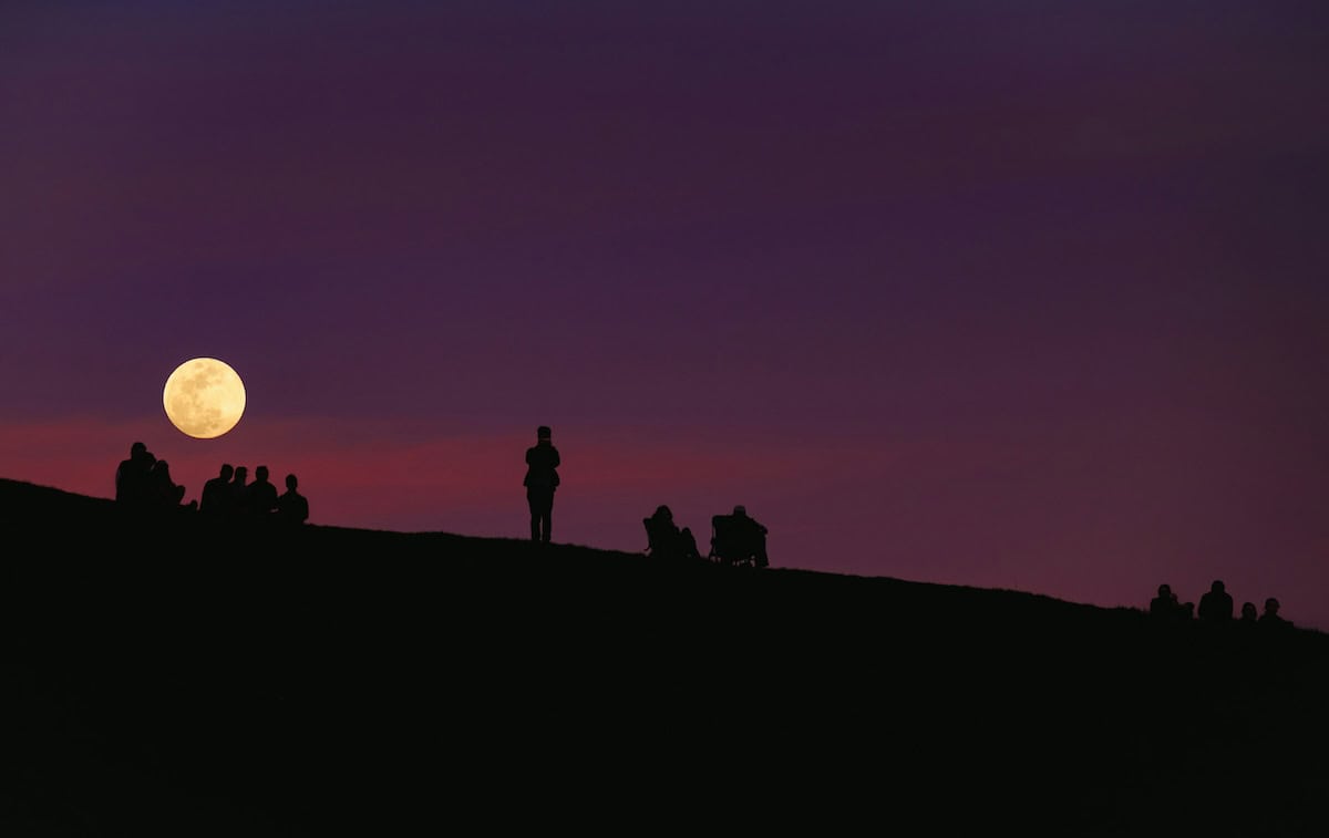 People watching the full moon from a hill at dusk.