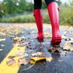 Autumn fall concept with colorful leaves and rain boots outside. Close up of woman feet walking in red boots.