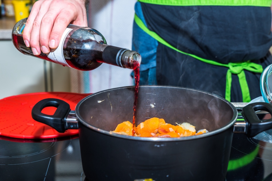 Male hand holding bottle and pouring red wine into pan casserole with frying carrots and onions on modern electric cooker.