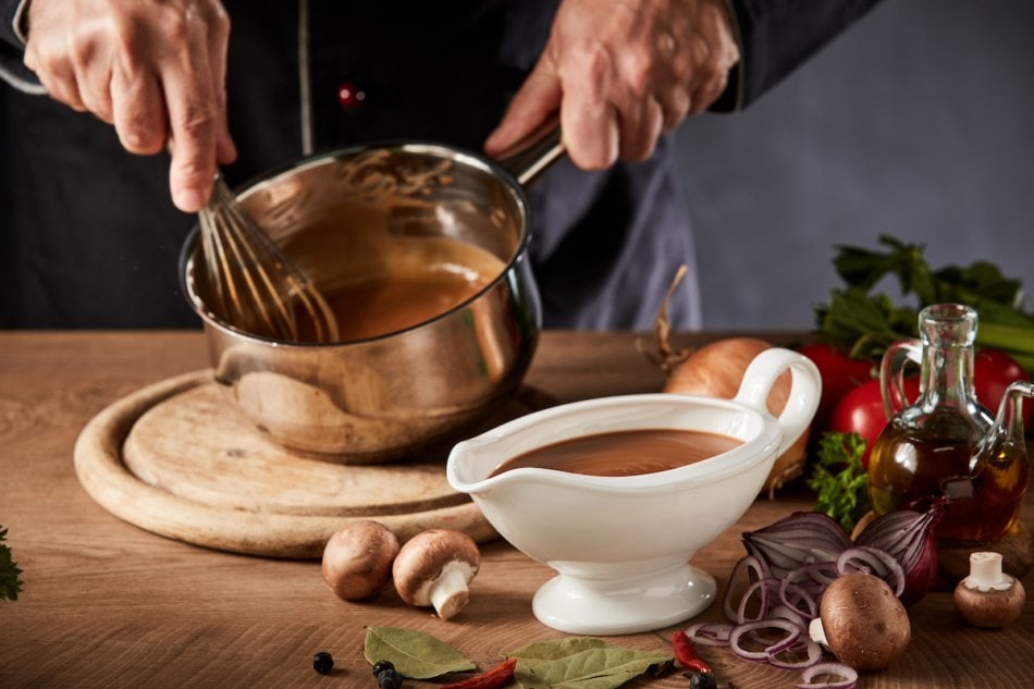 Chef whisking up a rich spicy gravy for dinner in a close up on a full sauce boat surrounded by ingredients as he whisks the mixture in a pot behind.