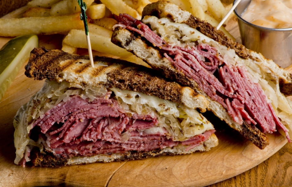 Reuben Sandwich, classic traditional American bar/pub menu item, on grilled rye bread, corned beef, Swiss cheese, sauerkraut and topped with thousand island dressing and french fries in background.