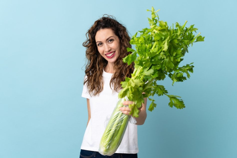Young woman holding a celery isolated on blue background with happy expression.