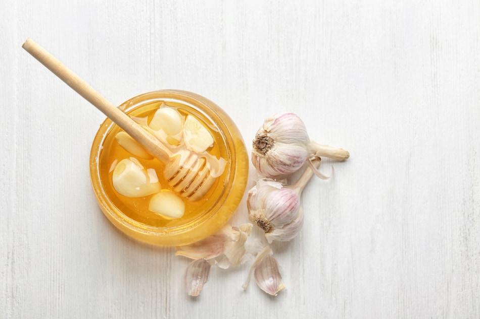 Glass jar with honey and garlic on white wooden background, top view
