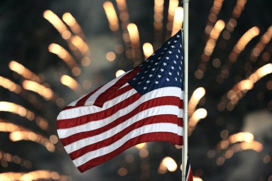 The Stars and Stripes wave through the night sky with fireworks in the background.