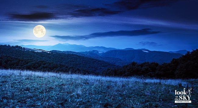 May night sky with the full Flower Moon over blue mountains.