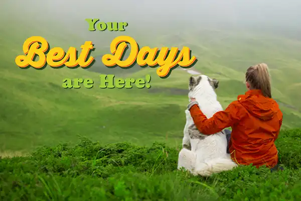 Woman in a red coat with her arm around a dog looking out over a misty green field.
