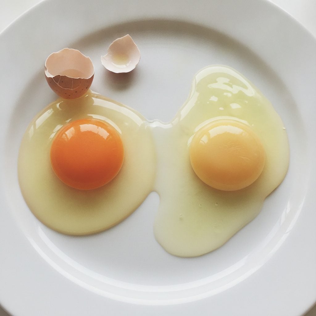 Two raw eggs cracked side by side onto a white plate showing the difference between a fresh tall-yolk egg and an older flat-spread egg.