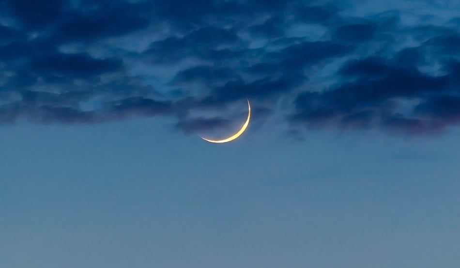 Crescent moon shining through dark, cloudy sky at dusk.