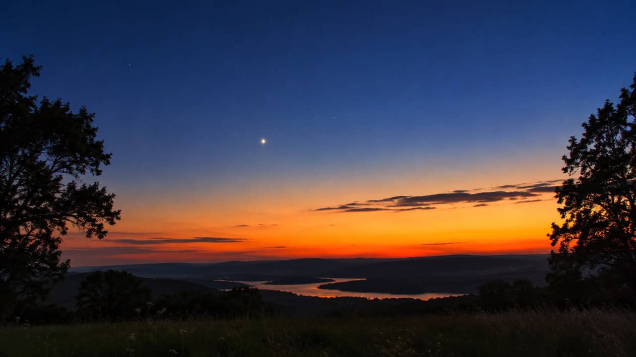 Venus visible after sunset over a scenic landscape with trees and water.