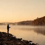 An angler casting from a rocky shore at dawn, mist on the water and autumn colour on the far bank, with a pale moon low in the sky.