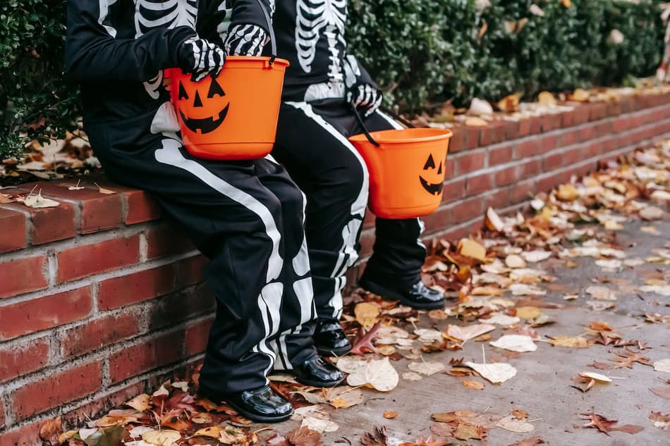 Children in skeleton costumes sitting on a brick wall with pumpkin buckets for trick-or-treating.