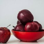 Red apples in a bowl with one apple outside on a white surface.