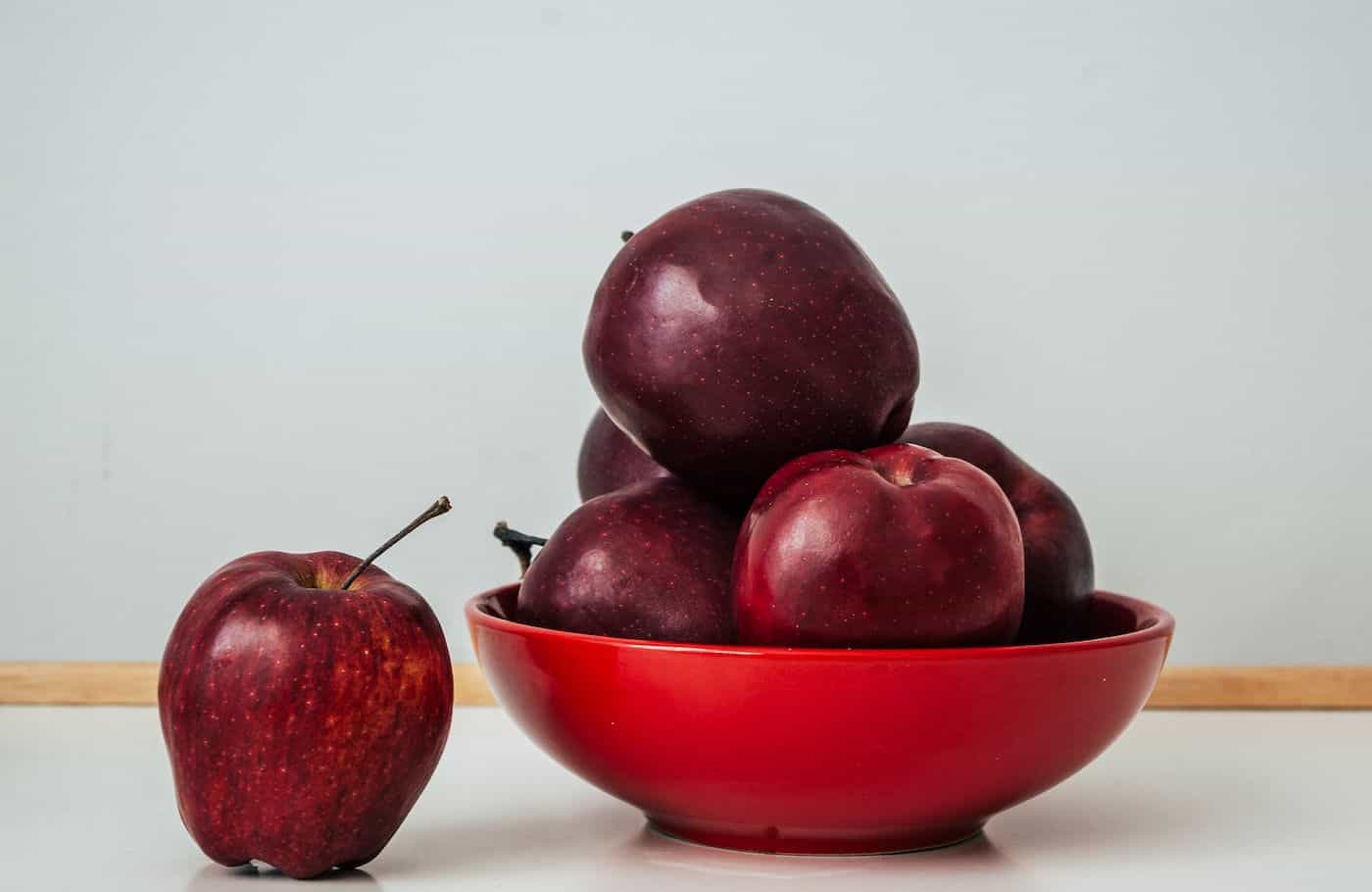 Red apples in a bowl with one apple outside on a white surface.