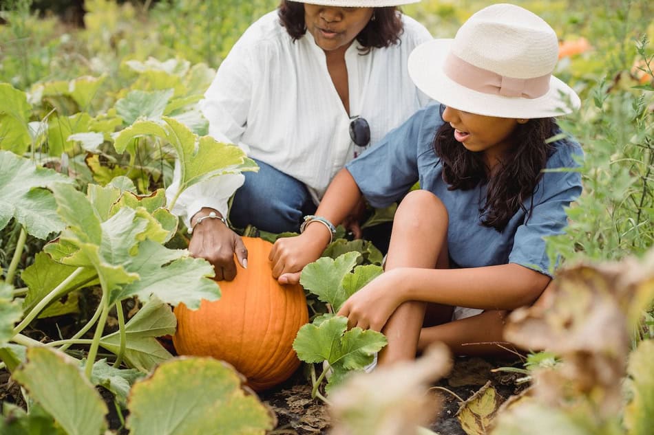 Two girls harvesting a pumpkin in a lush garden during fall.