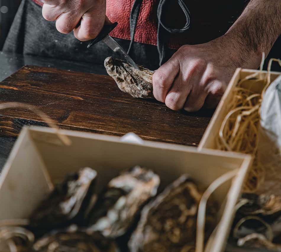 Person shucking oysters on a wooden table with a knife and oyster shell.