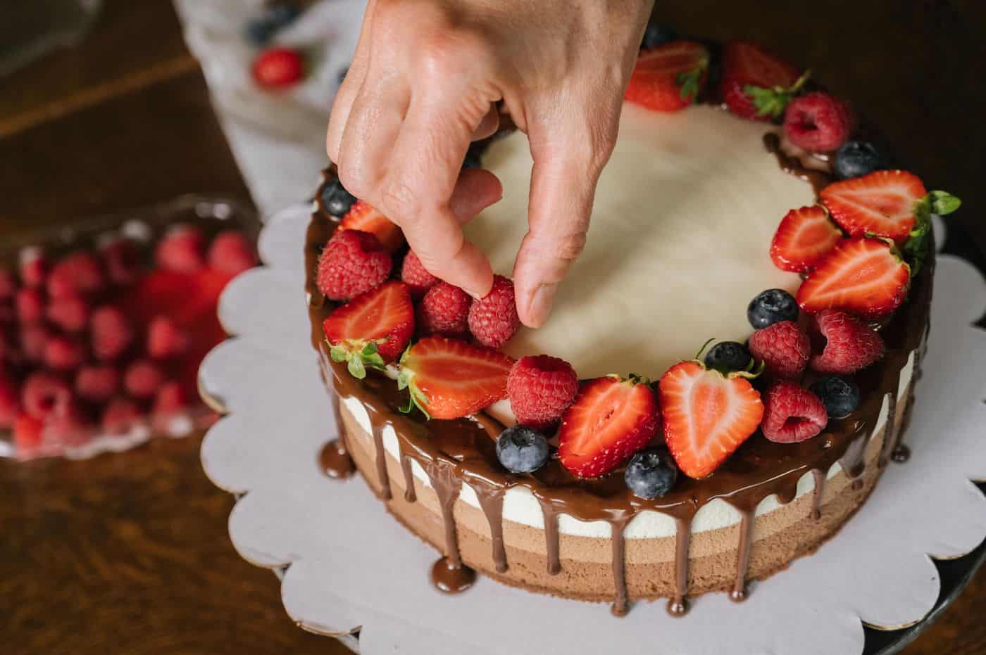 Hand placing fresh berries on a chocolate cake with strawberries and blueberries.